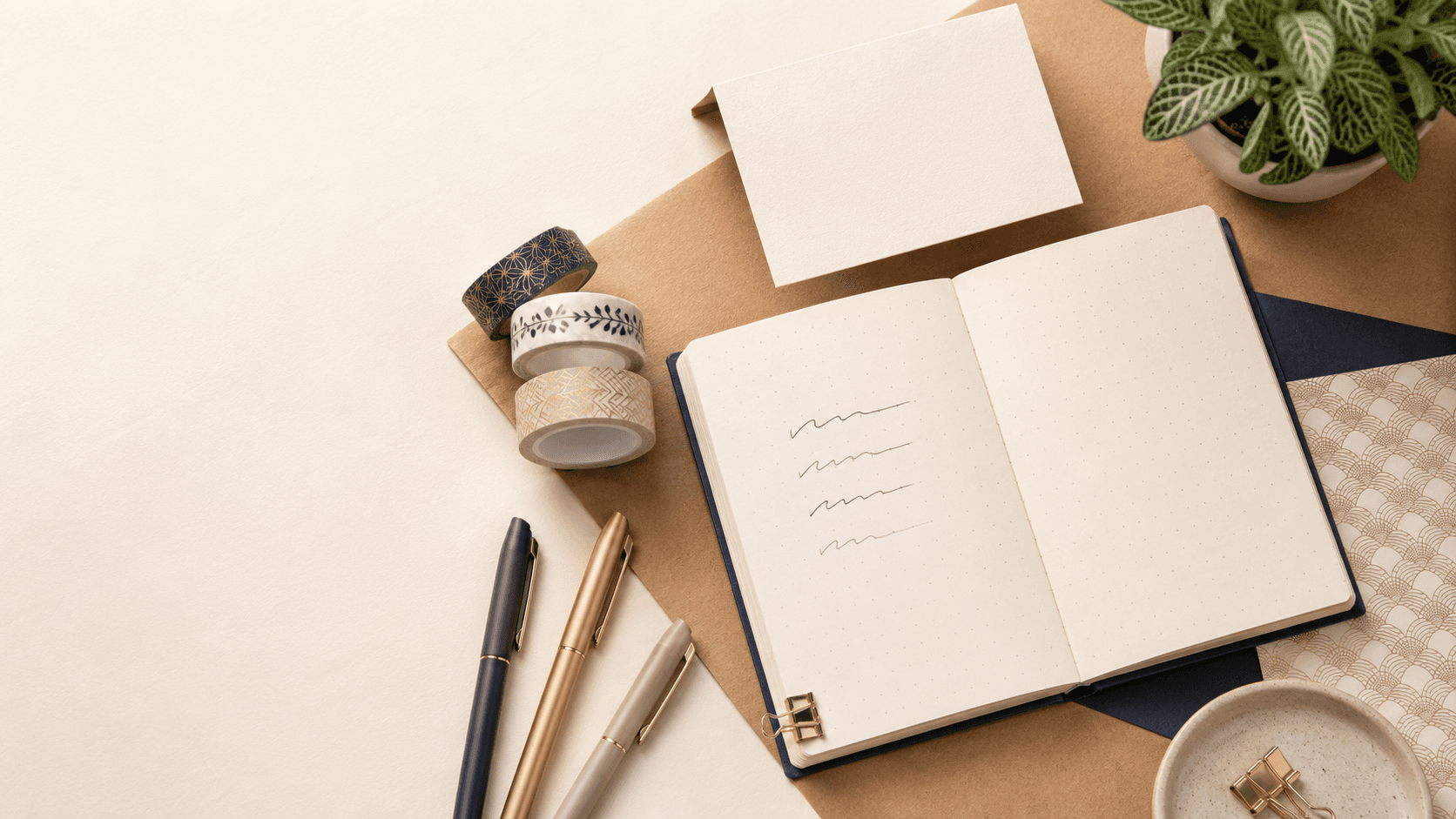An open dotted journal, pens, washi tape and a greeting card on a stationery desk
